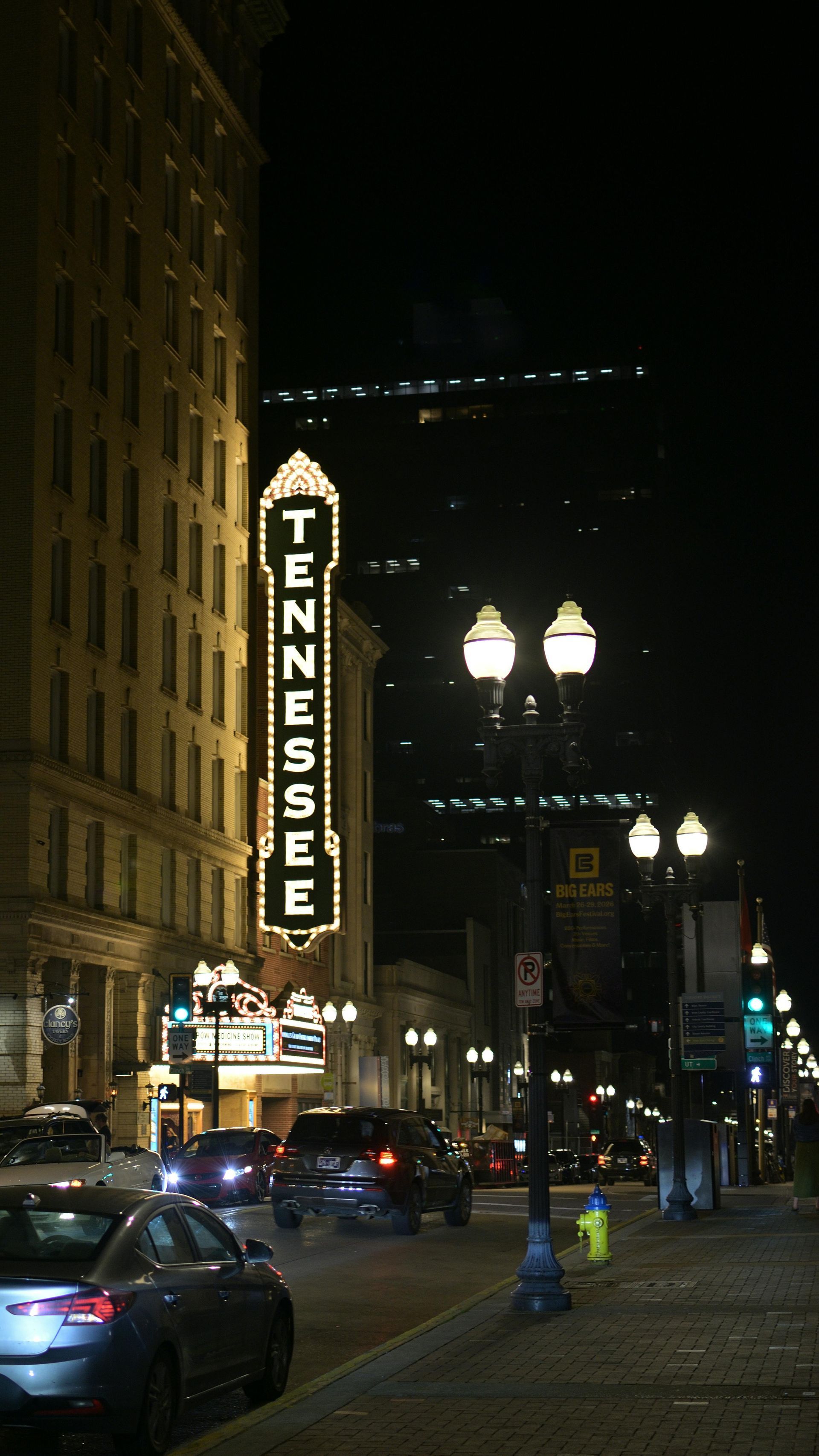 Night street scene with a lit Tennessee theater marquee, cars, and pedestrians on a city block.