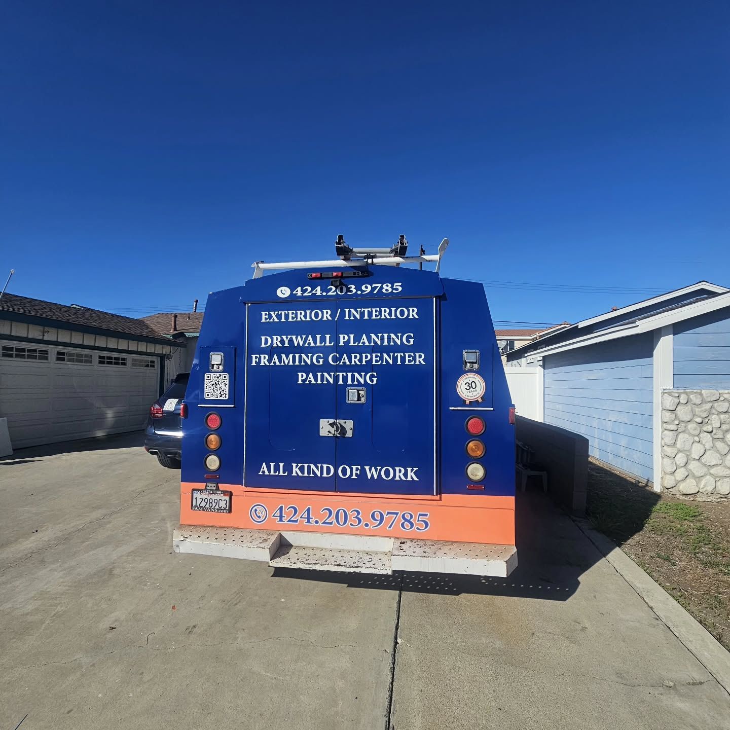 Blue work truck with business information on the back, parked on a driveway on a sunny day.