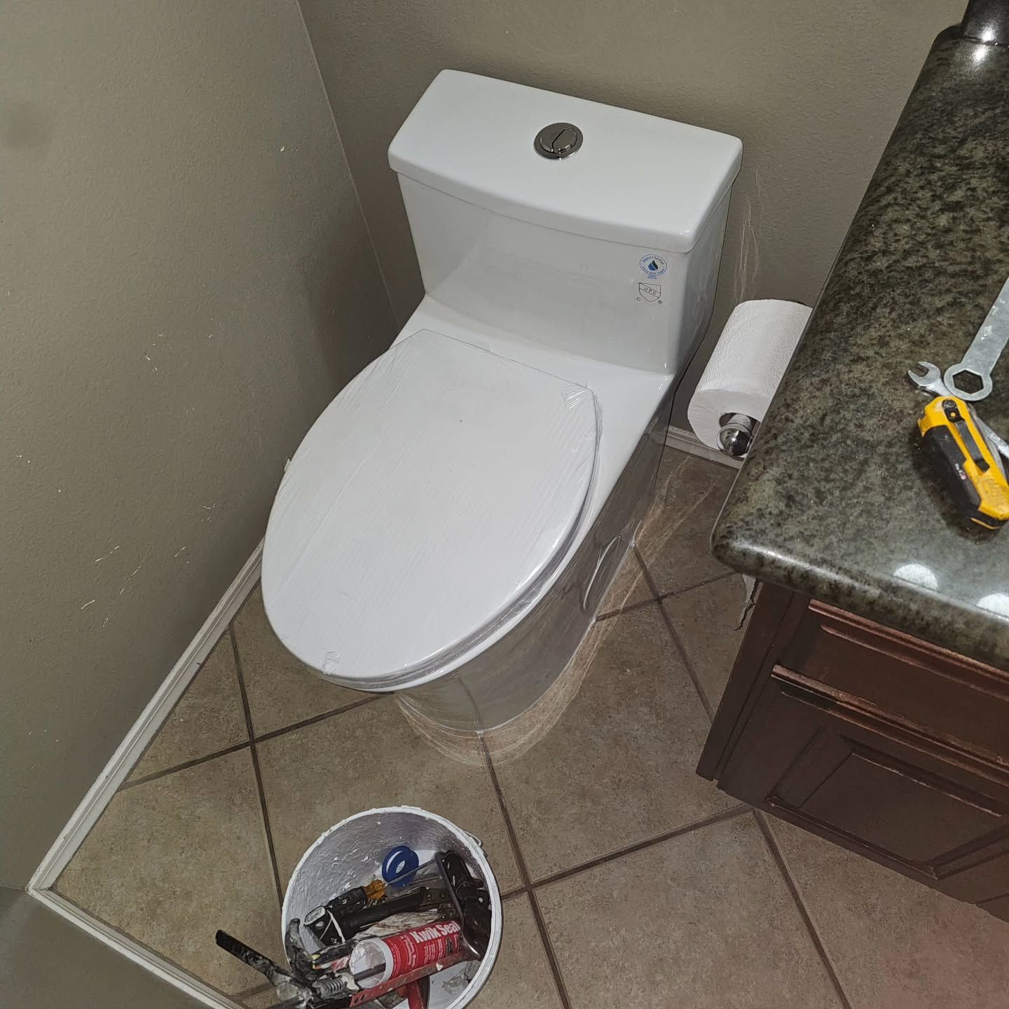 White toilet next to a dark countertop. A bucket of tools sits on the floor.