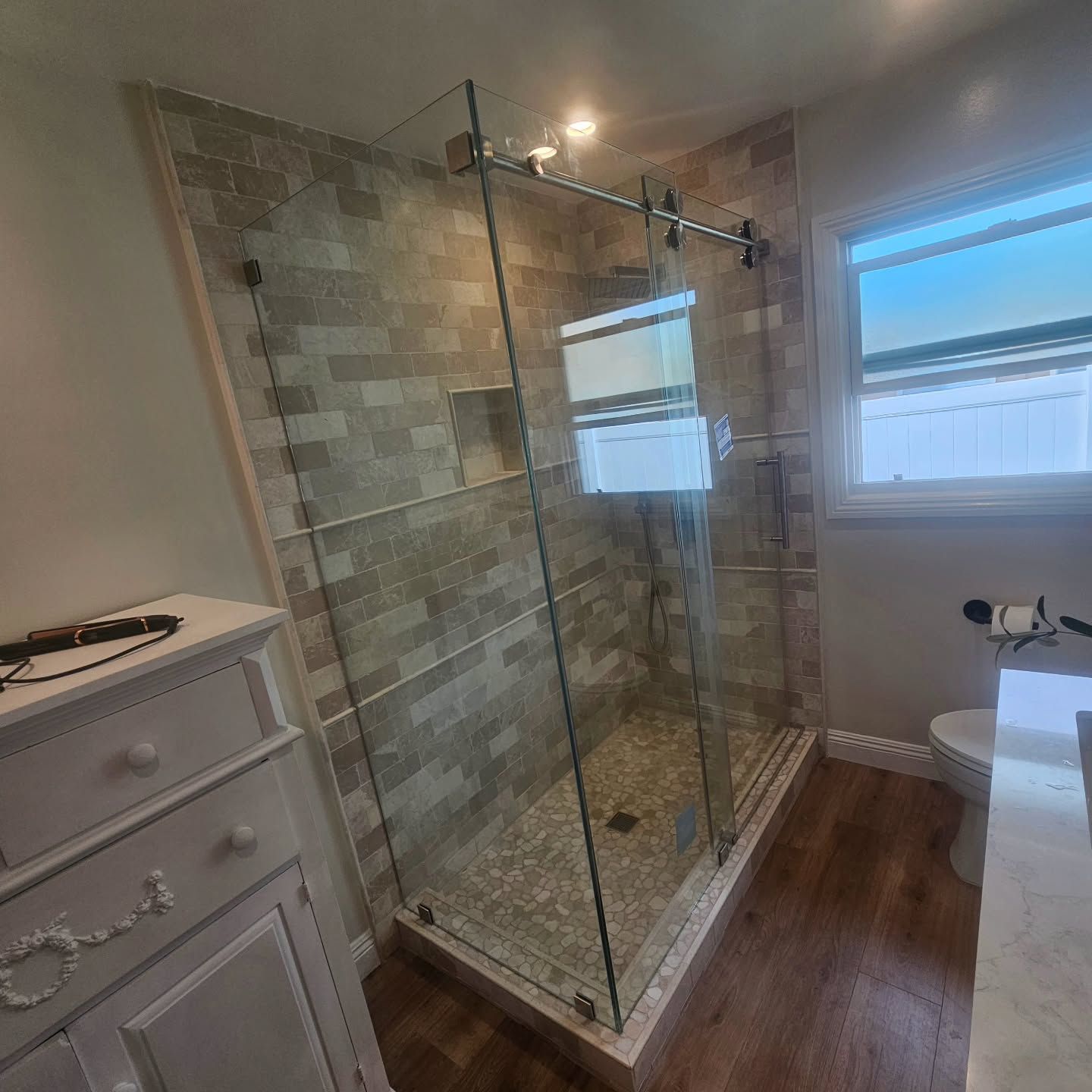 Bathroom with glass shower, tile walls, wood floor, white vanity, and toilet near a window.