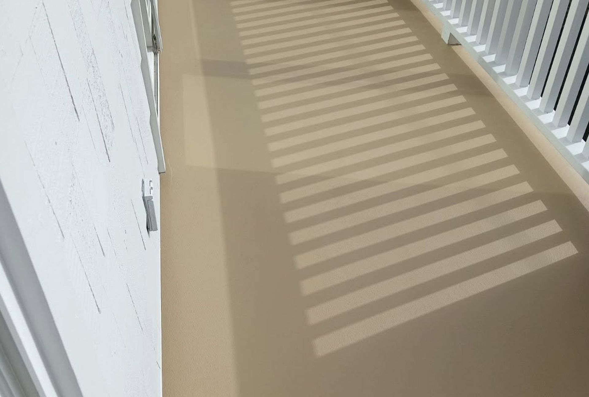 Shadow of a slatted object on a light brown surface with a white railing and wall.