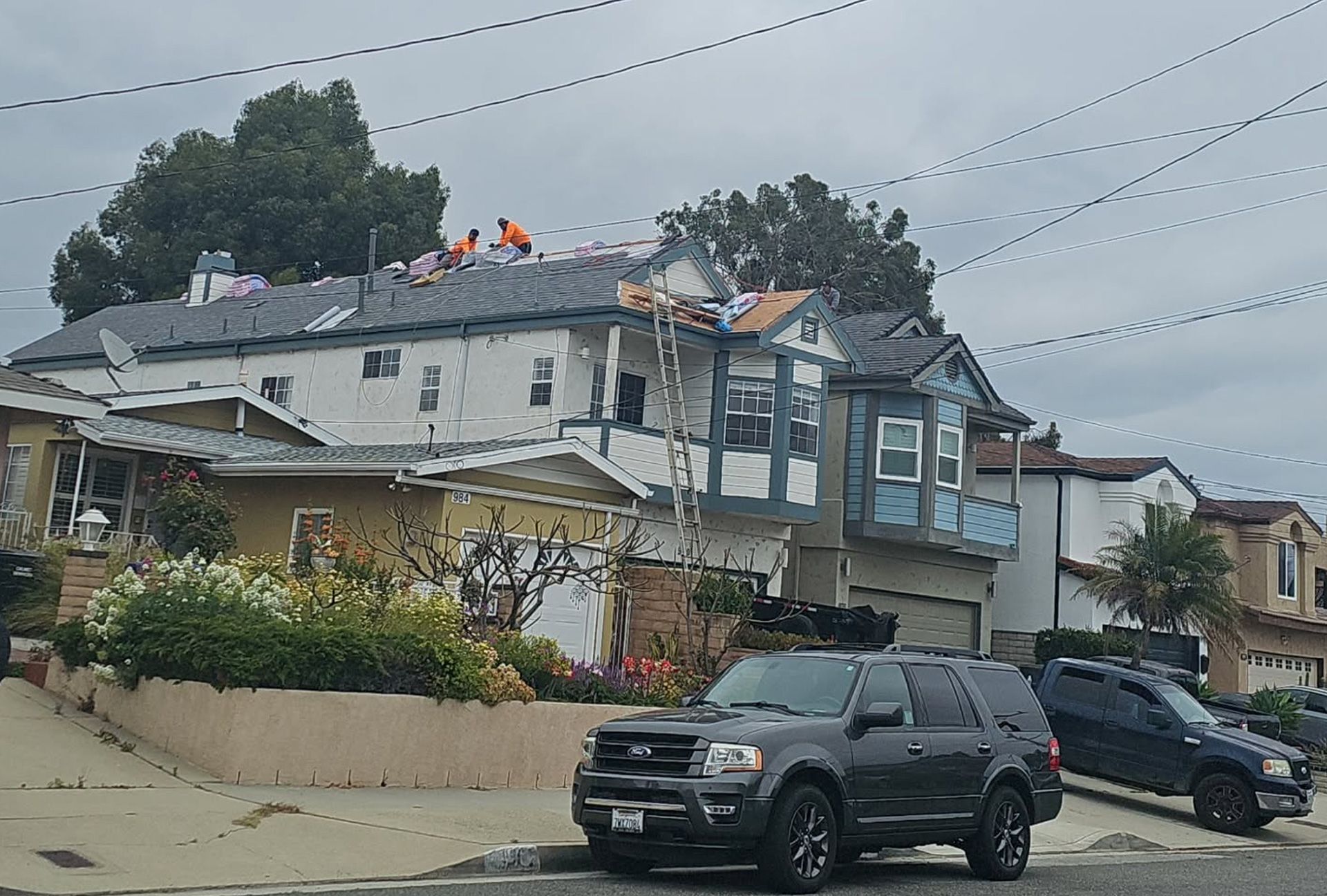 Workers on a house roof, removing and installing new shingles. Dark SUV parked in front.