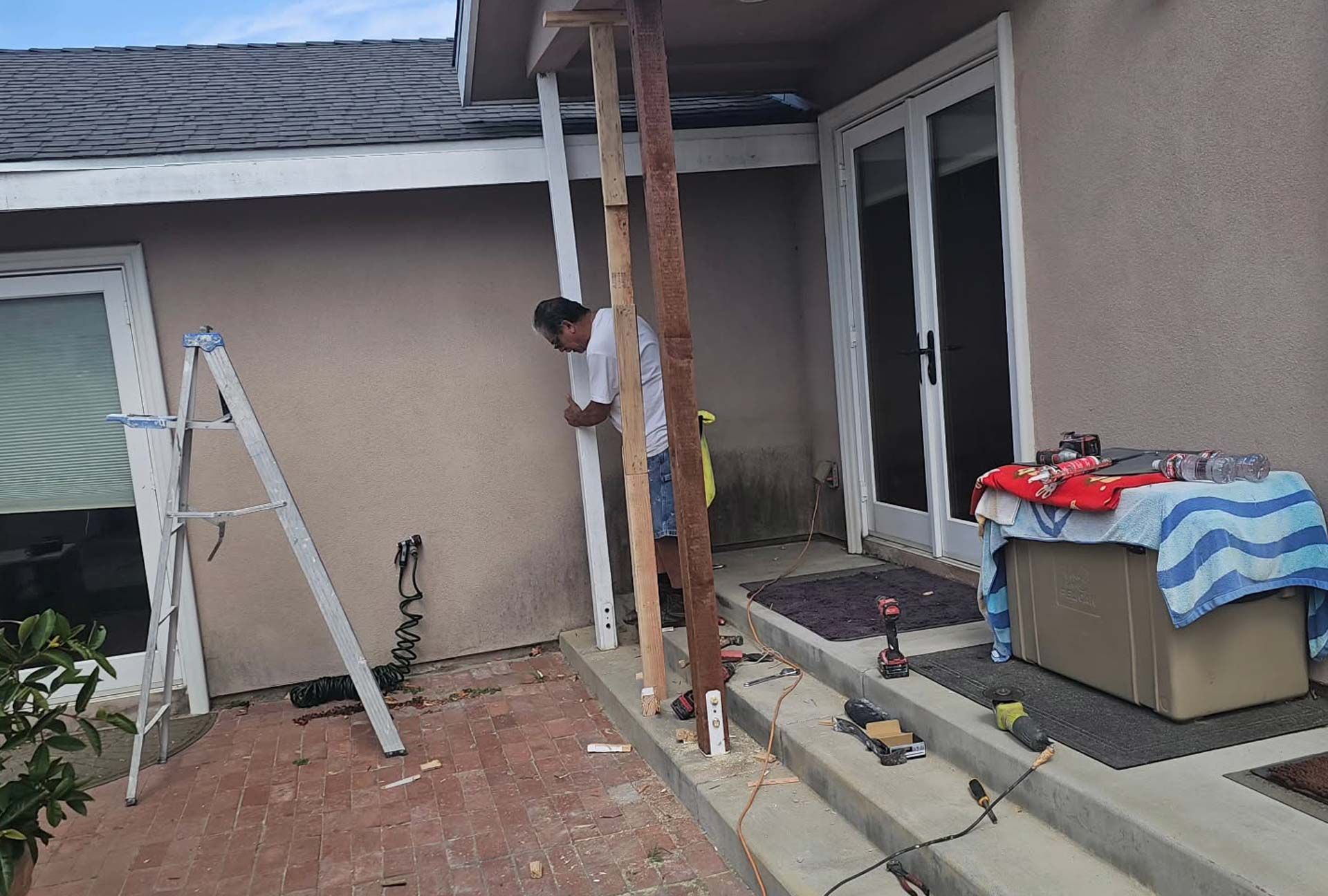 Person building a porch with wooden posts next to a house with white doors. Tools are on the ground.