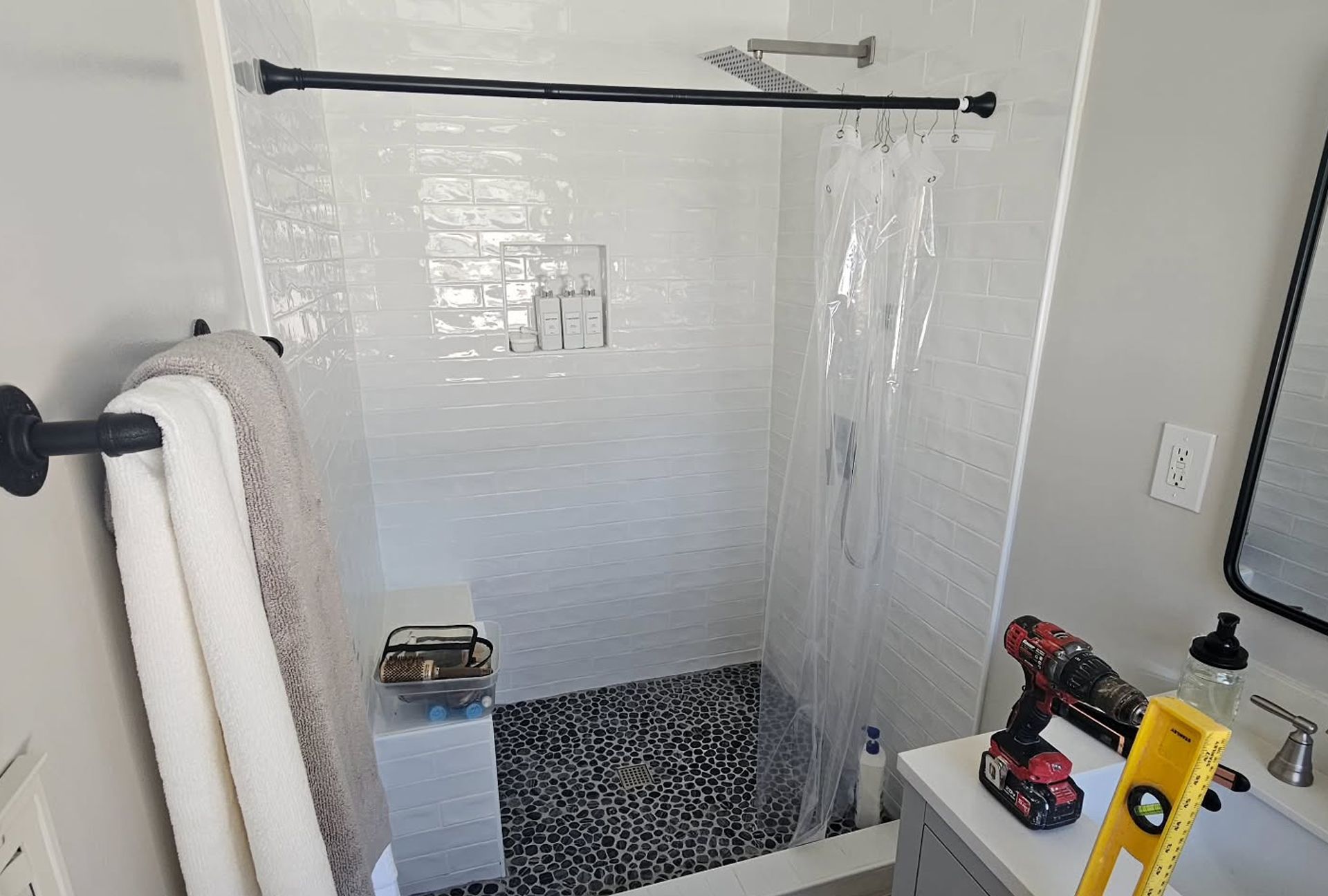 Bathroom with white tiled shower, black pebble floor, shower curtain, towels, and a vanity with a drill and level.