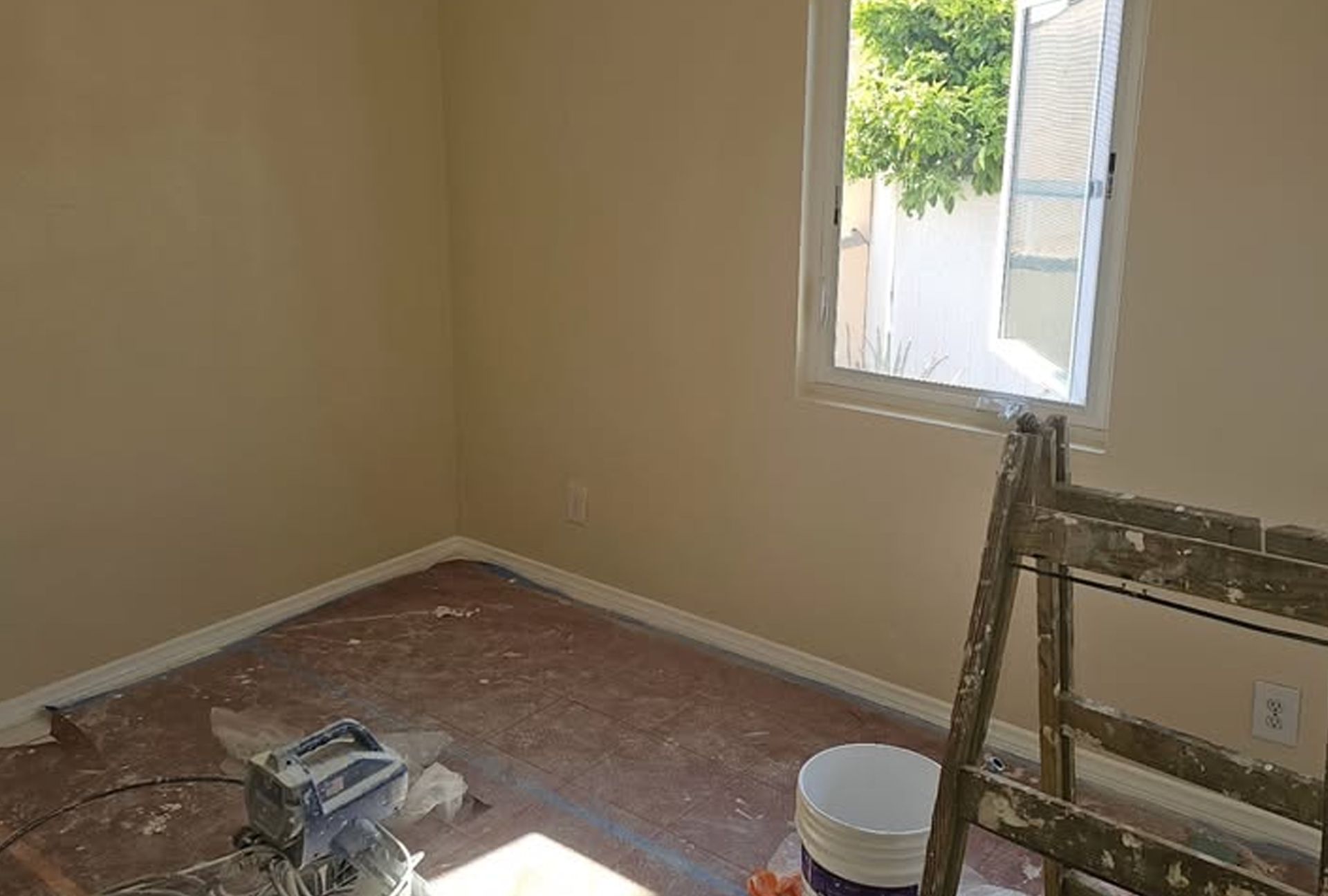 Room interior with tan walls, window, ladder, and paint bucket; construction in progress.