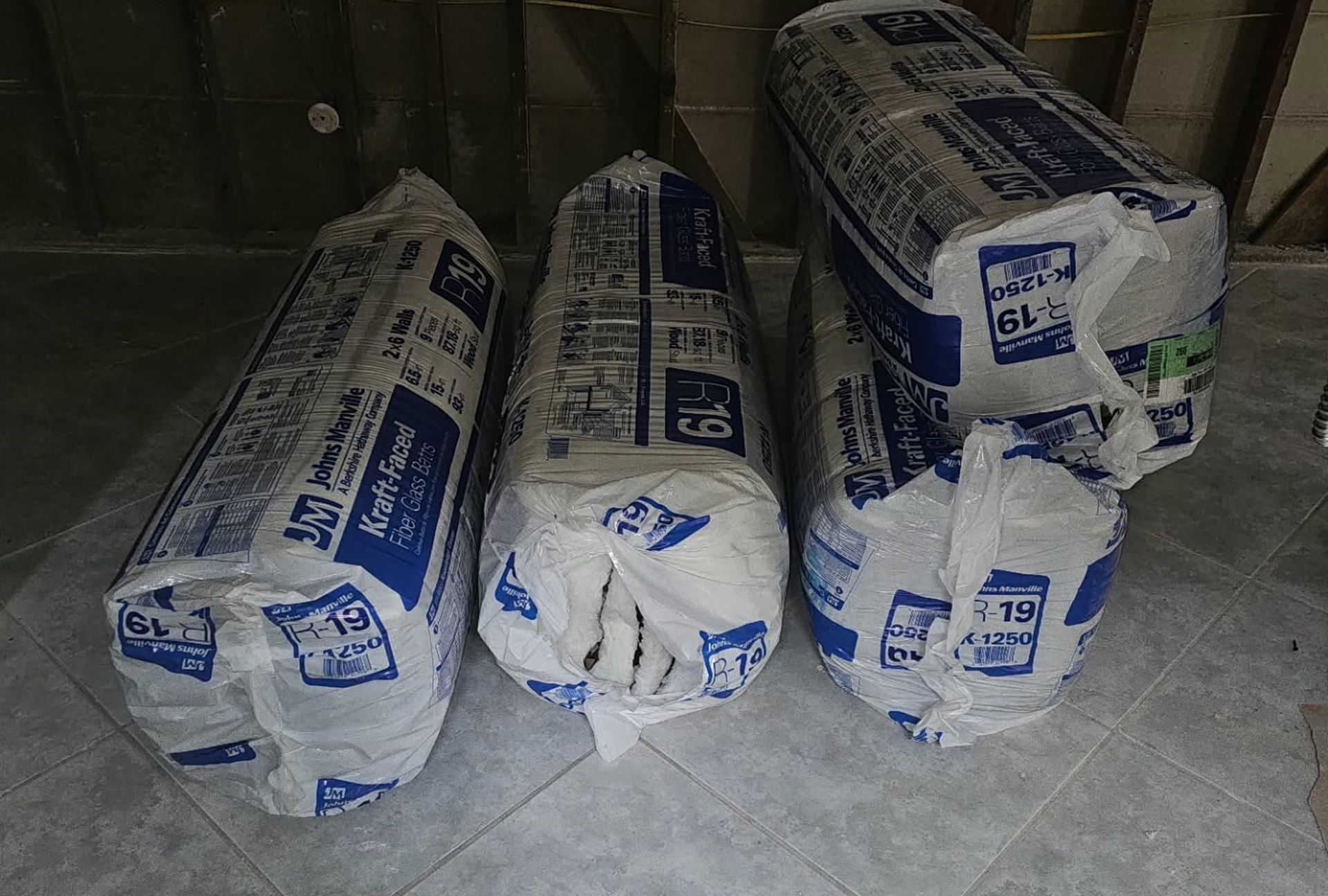 Four rolls of fiberglass insulation in plastic packaging on a concrete floor.