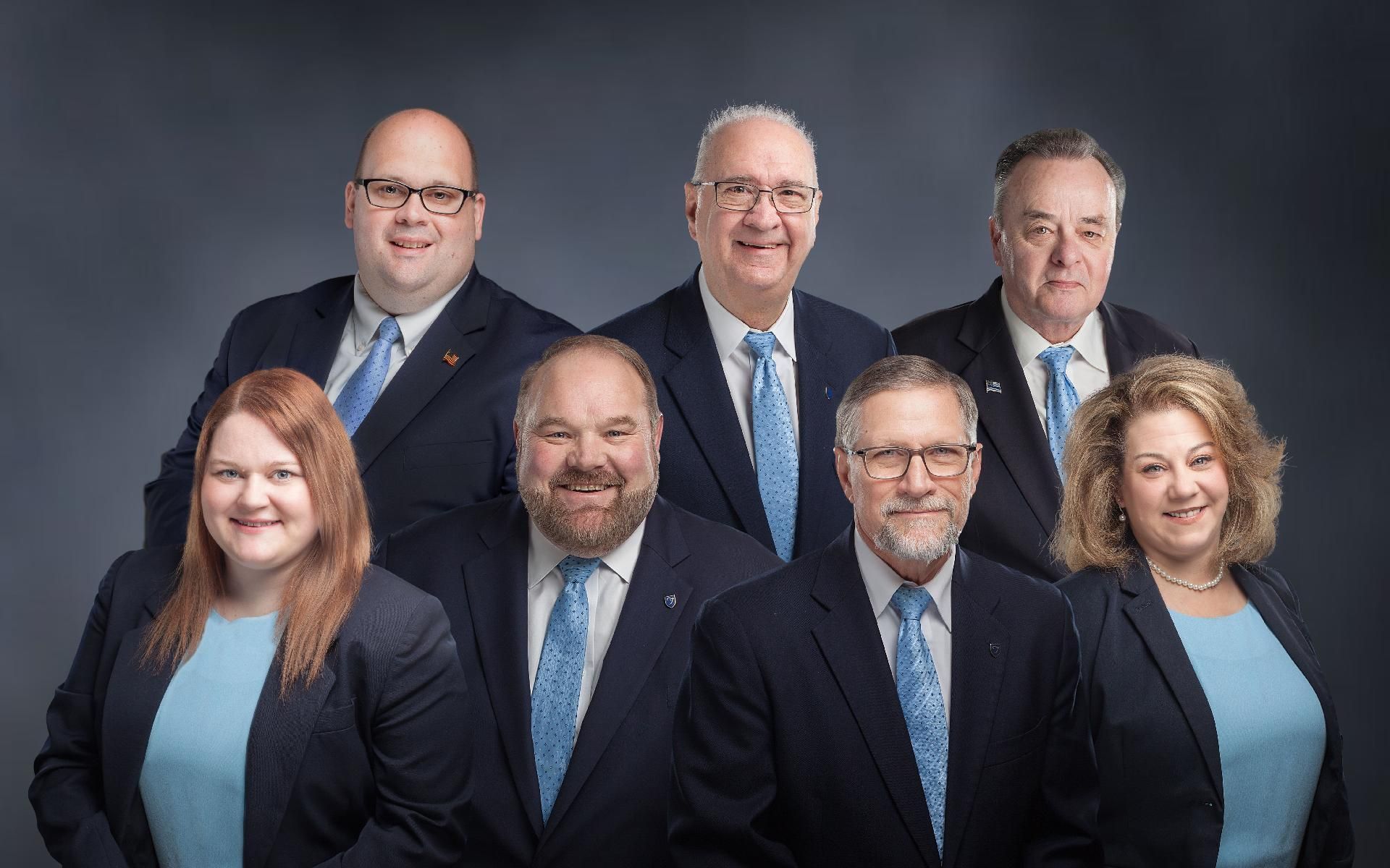 Seven professionally dressed people pose for a studio group portrait against a dark gray backdrop.