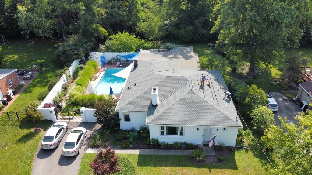Aerial view of a white house with a backyard pool, patio, and parked cars surrounded by trees and lawn