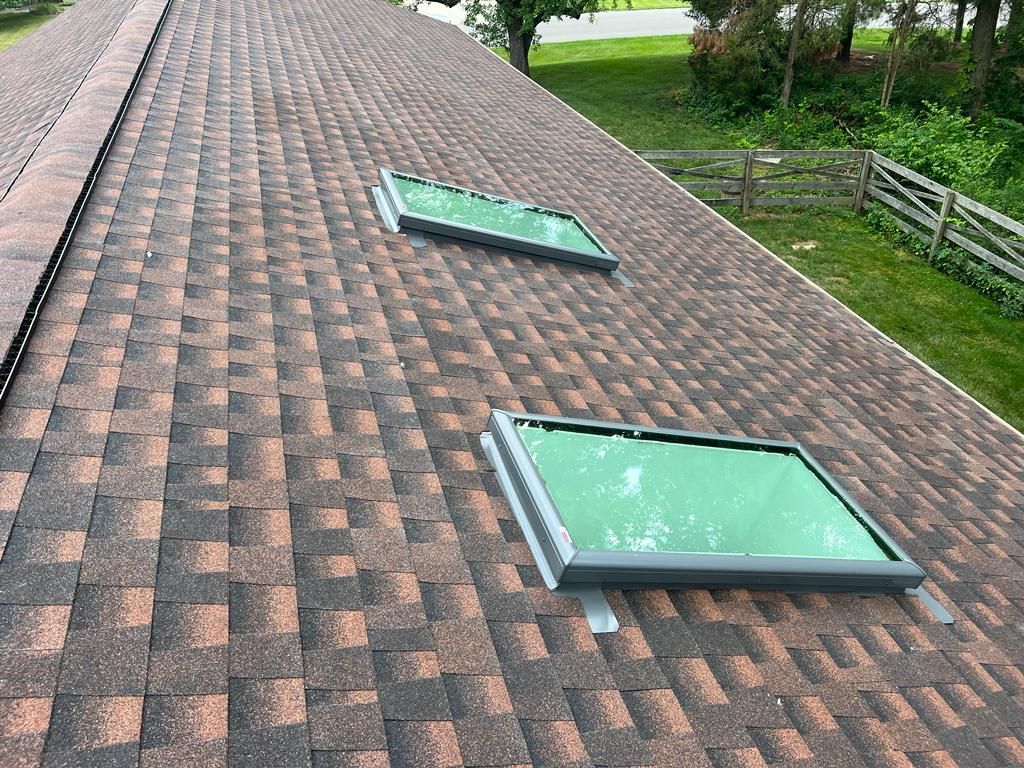 Brown shingled roof with two green skylights beside a grassy yard and fence
