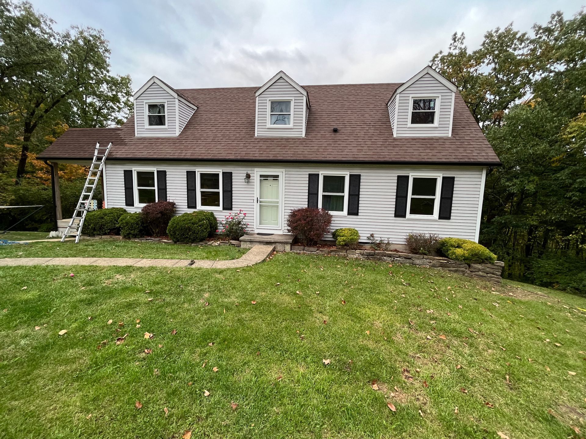 White house with dark roof, dormer windows, front steps, and a green lawn under cloudy skies