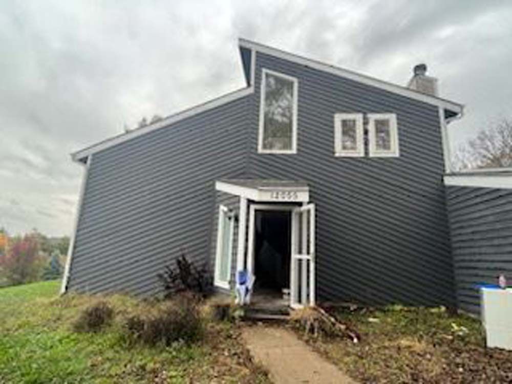 A large gray house with a white door and windows is sitting on top of a grassy hill.