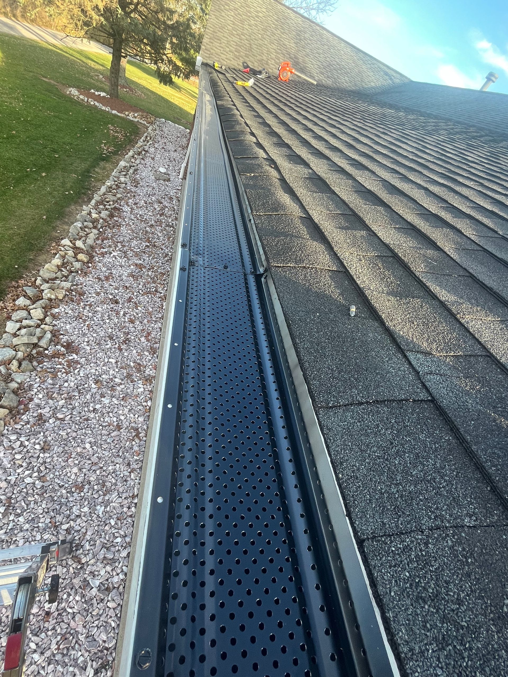 Roof gutter beside gray shingle roof, with gravel ground and grassy slope nearby.