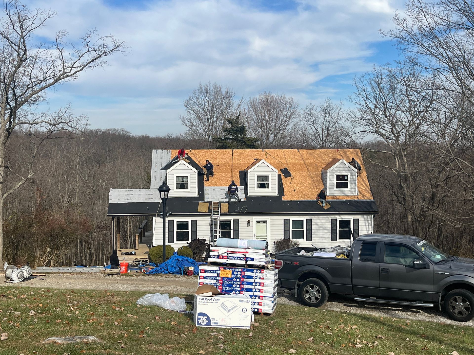 House under renovation with black pickup truck, stacked materials, and bare trees in a yard
