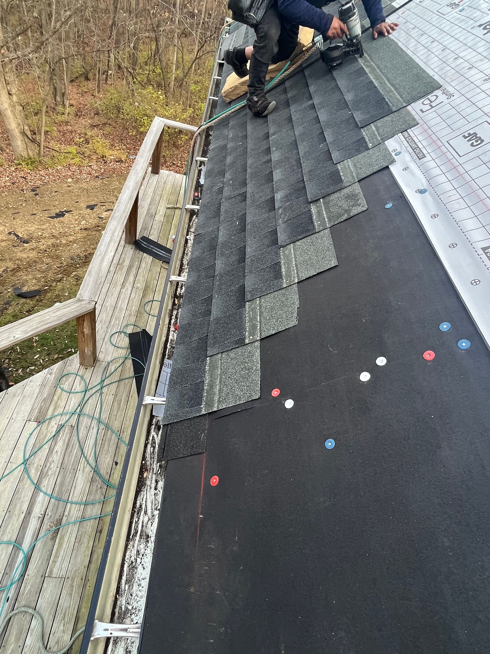 Worker installing dark shingles on a steep roof beside a railing and white roof edge.
