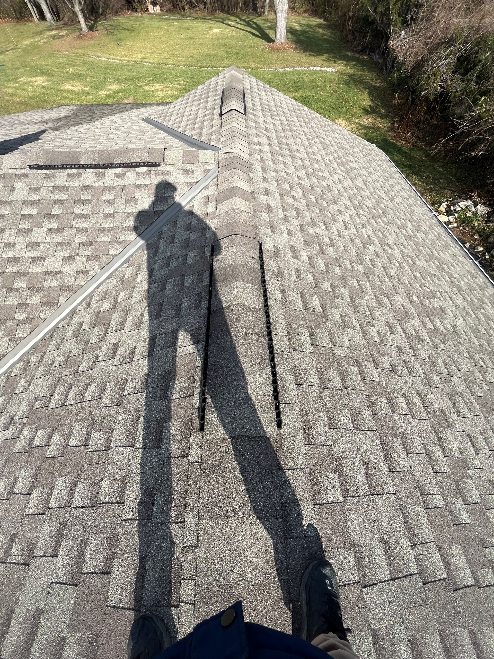 Person’s shadow on a textured gray roof beside a metal handrail, with grass and trees in the background