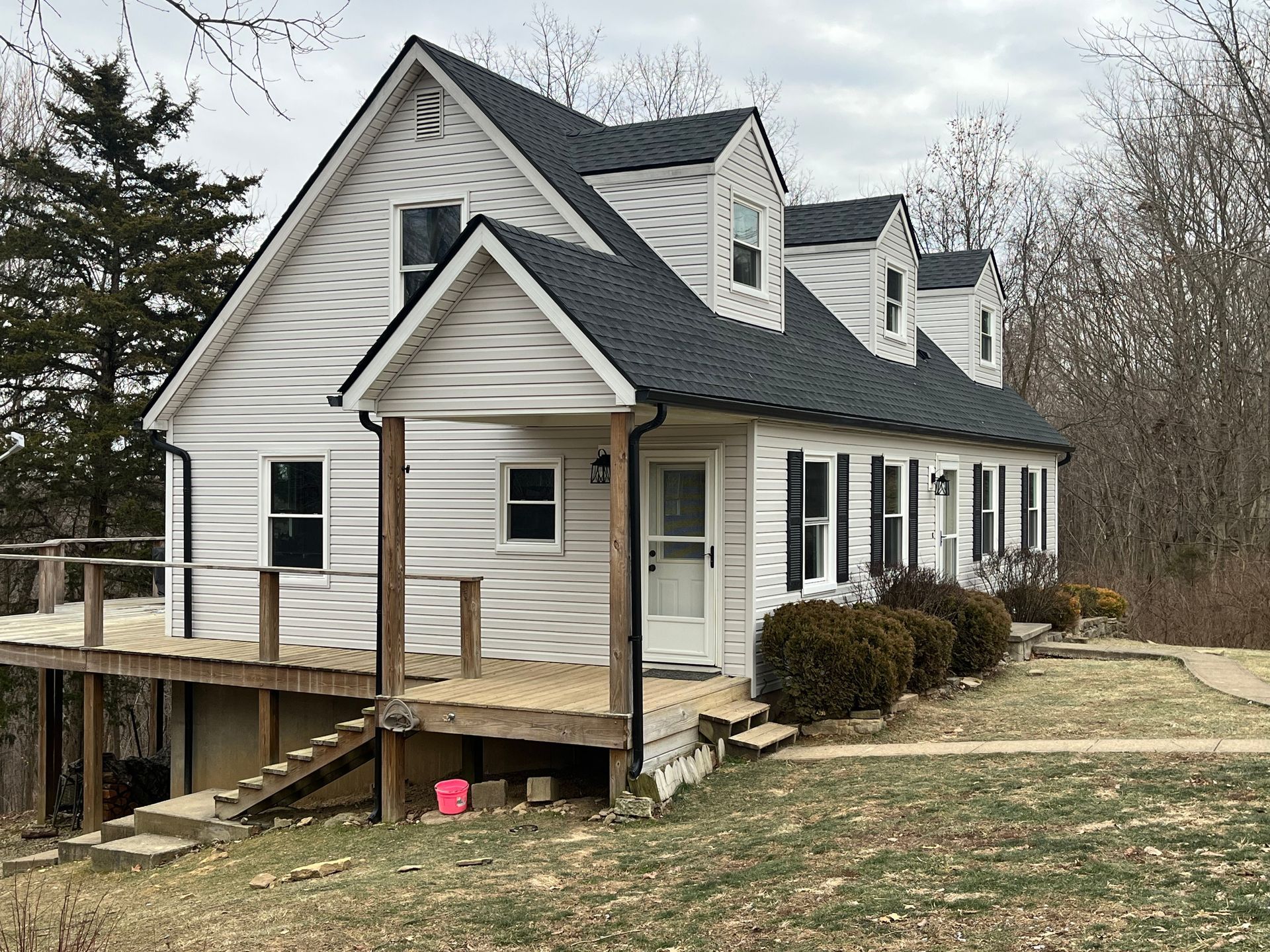 White house with dark roof and porch on a grassy lot, surrounded by leafless trees