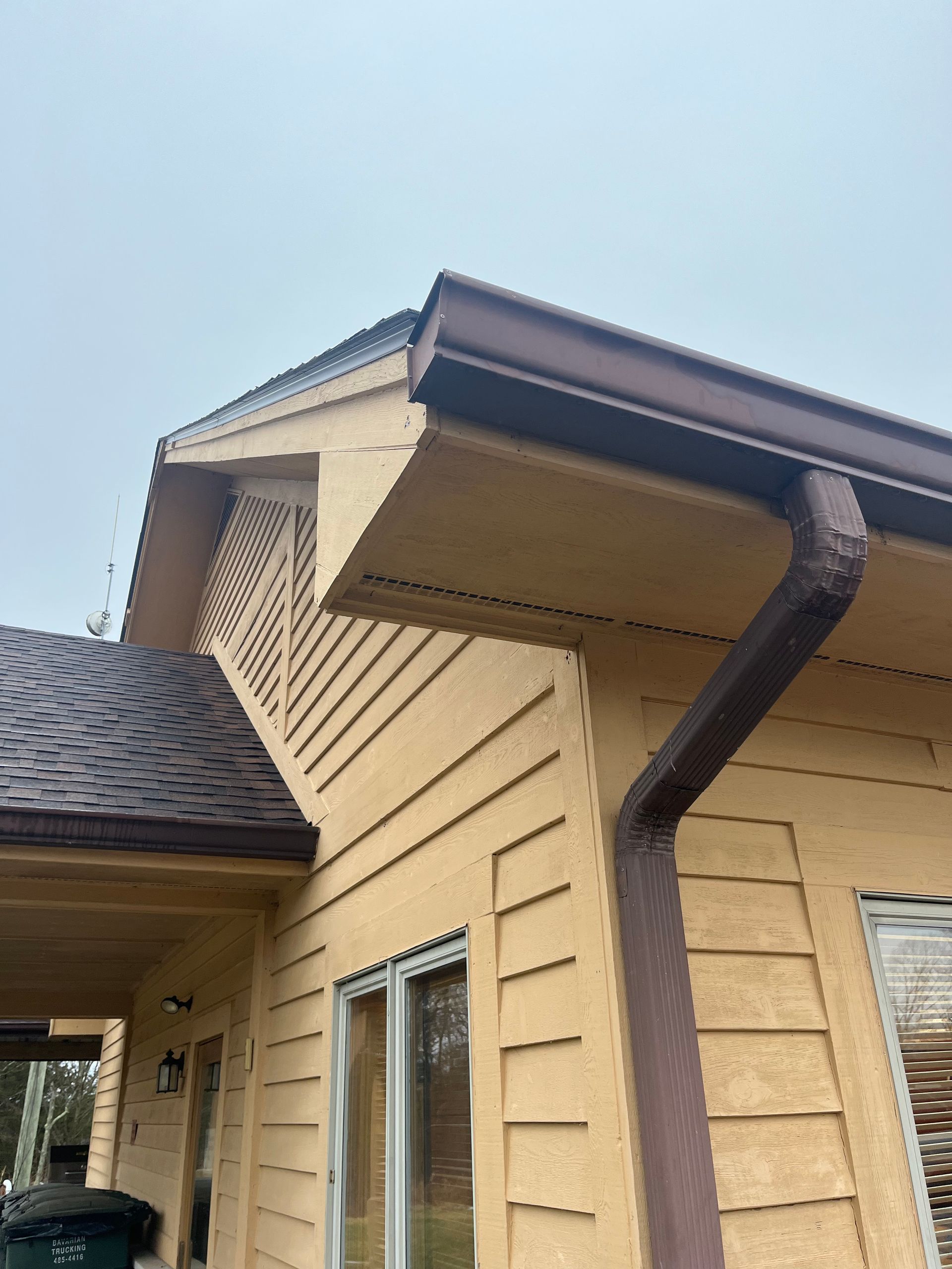 Close-up of a beige house exterior with roof eaves and a brown rain gutter against a blue sky