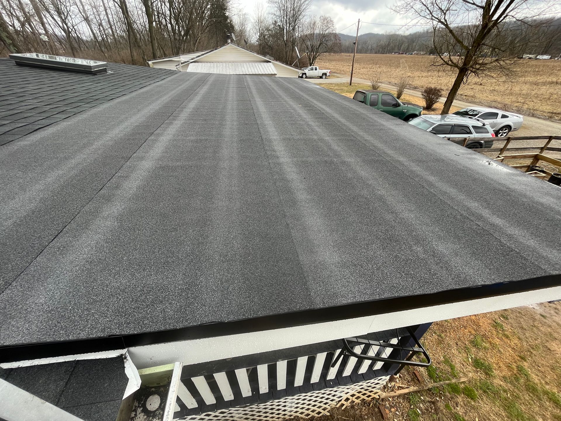 Roof with fresh black shingles overlooking a rural yard and parked cars on a cloudy day