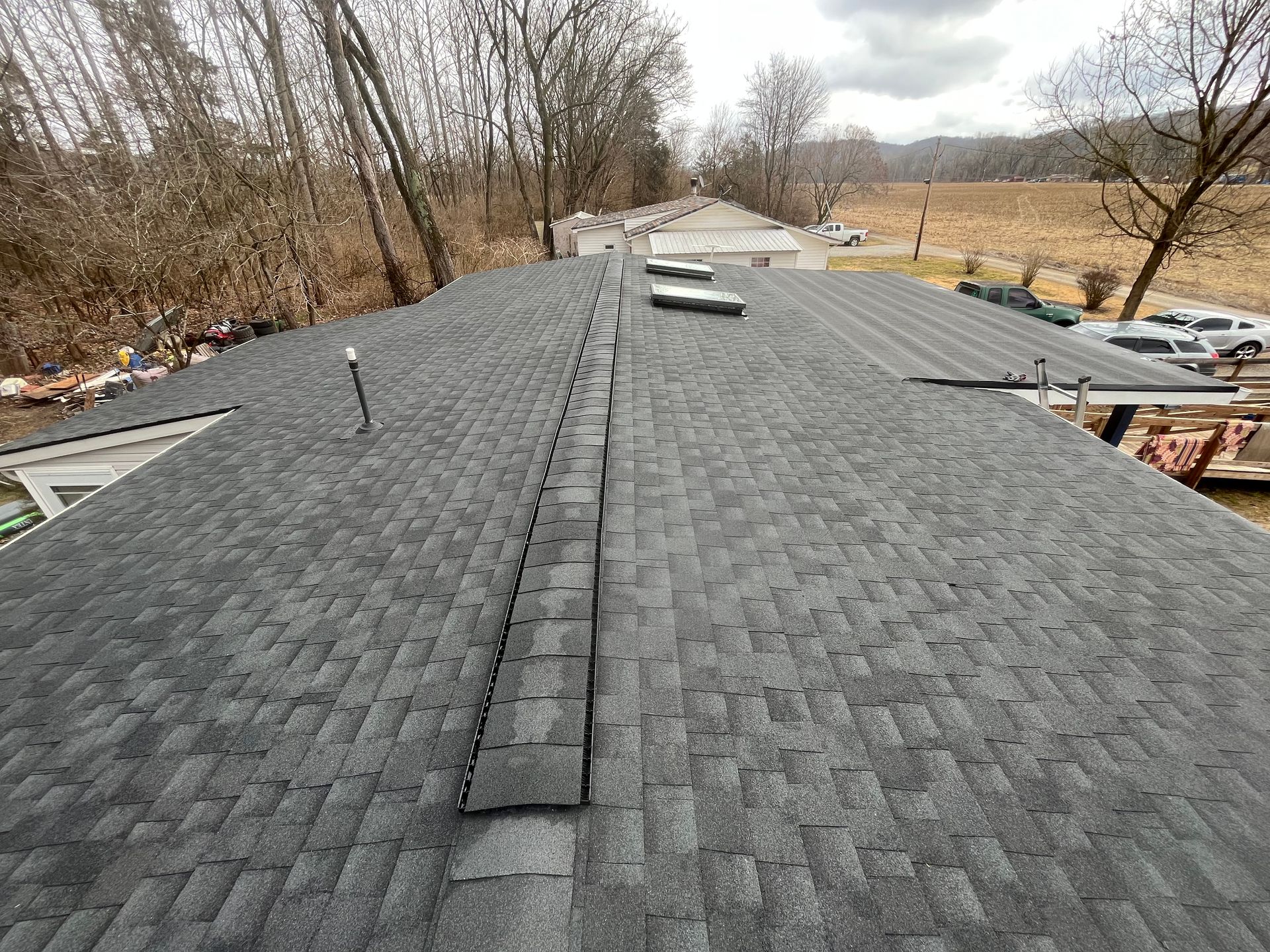 Roof under construction with dark shingles, a central ridge vent, and trees in the background.