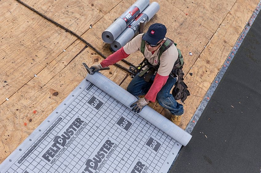 Worker installing roofing underlayment on a sloped roof, rolling out gray membrane near the edge.