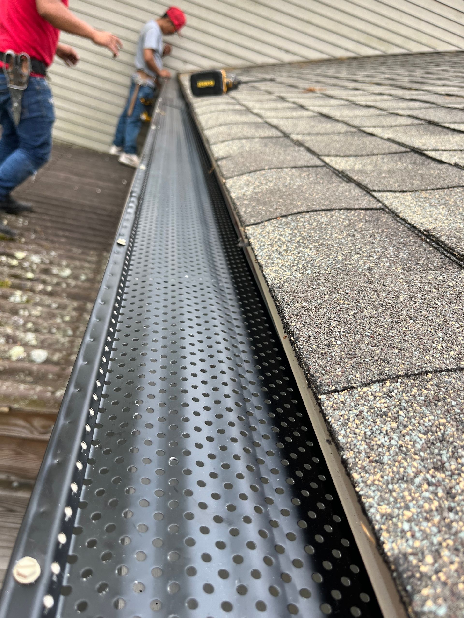 Workers installing a black gutter guard along a roof edge on a shingled roof