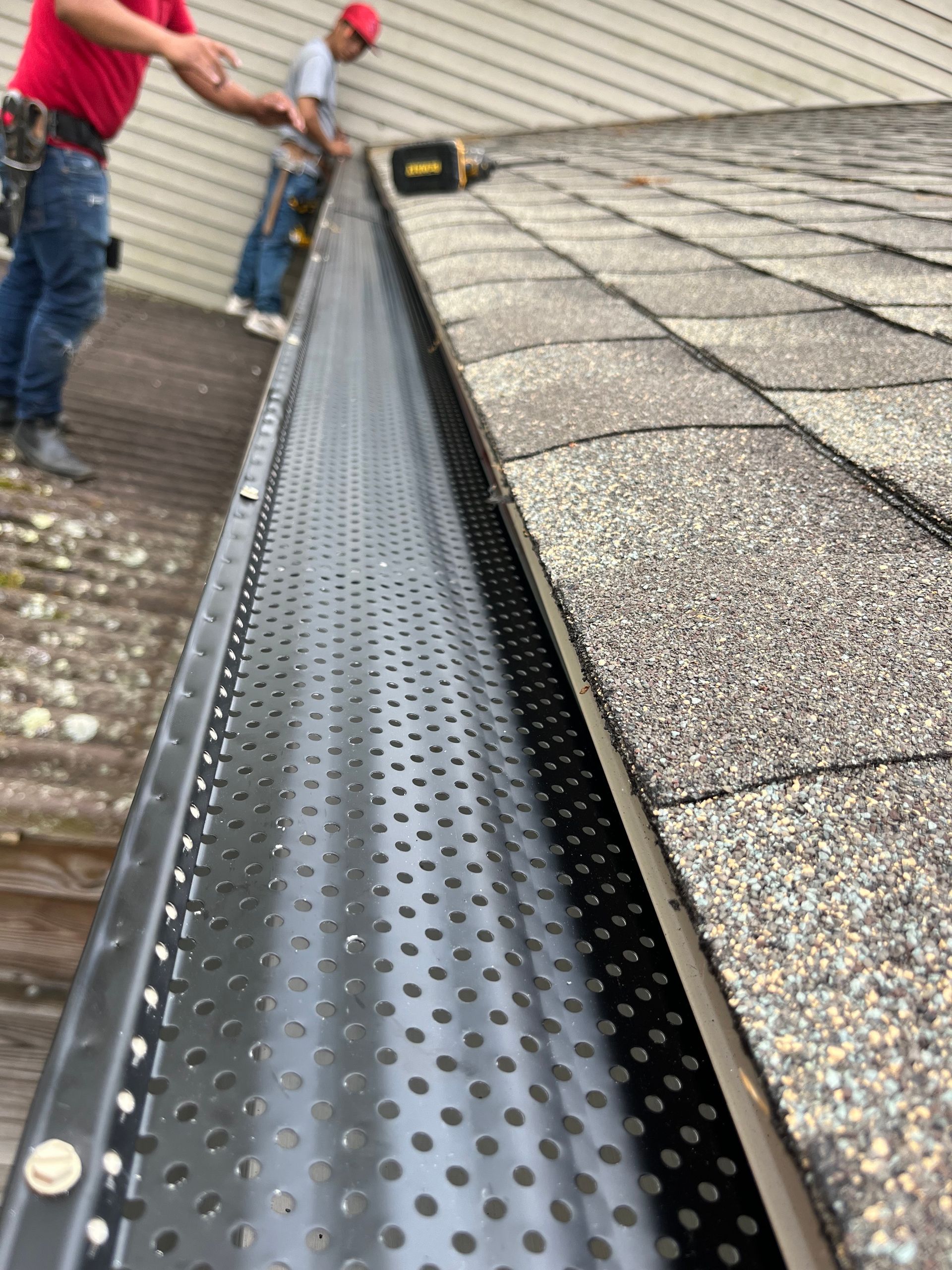 Perforated black roof gutter installed along a shingled roof edge, with workers on the roof.