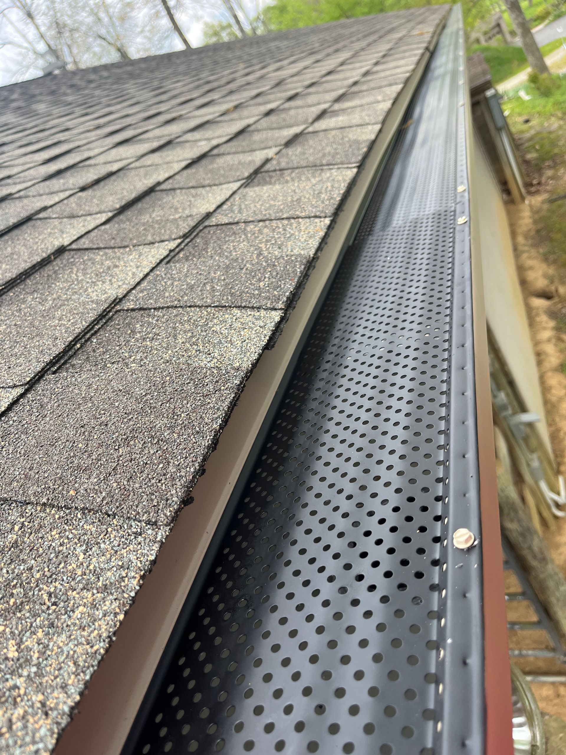 Gutter guard along shingled roof edge beside brick wall and tree branches