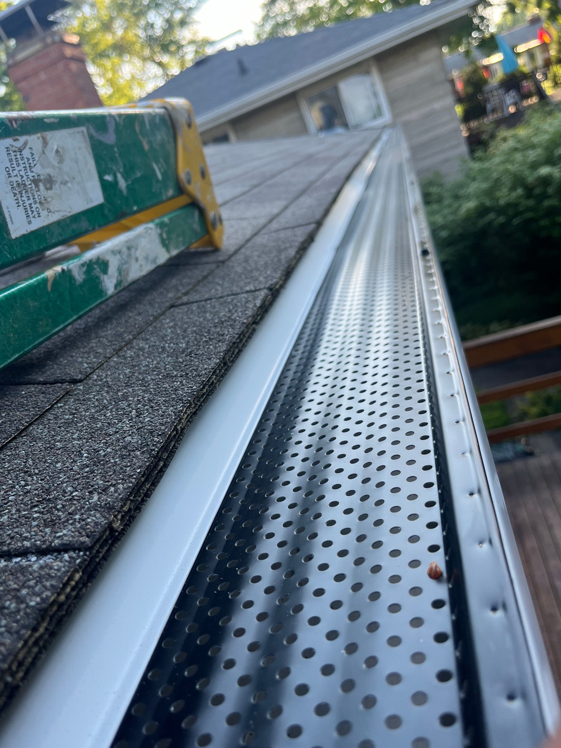 Metal roof gutter with perforated leaf guard along a shingled roof edge in daylight
