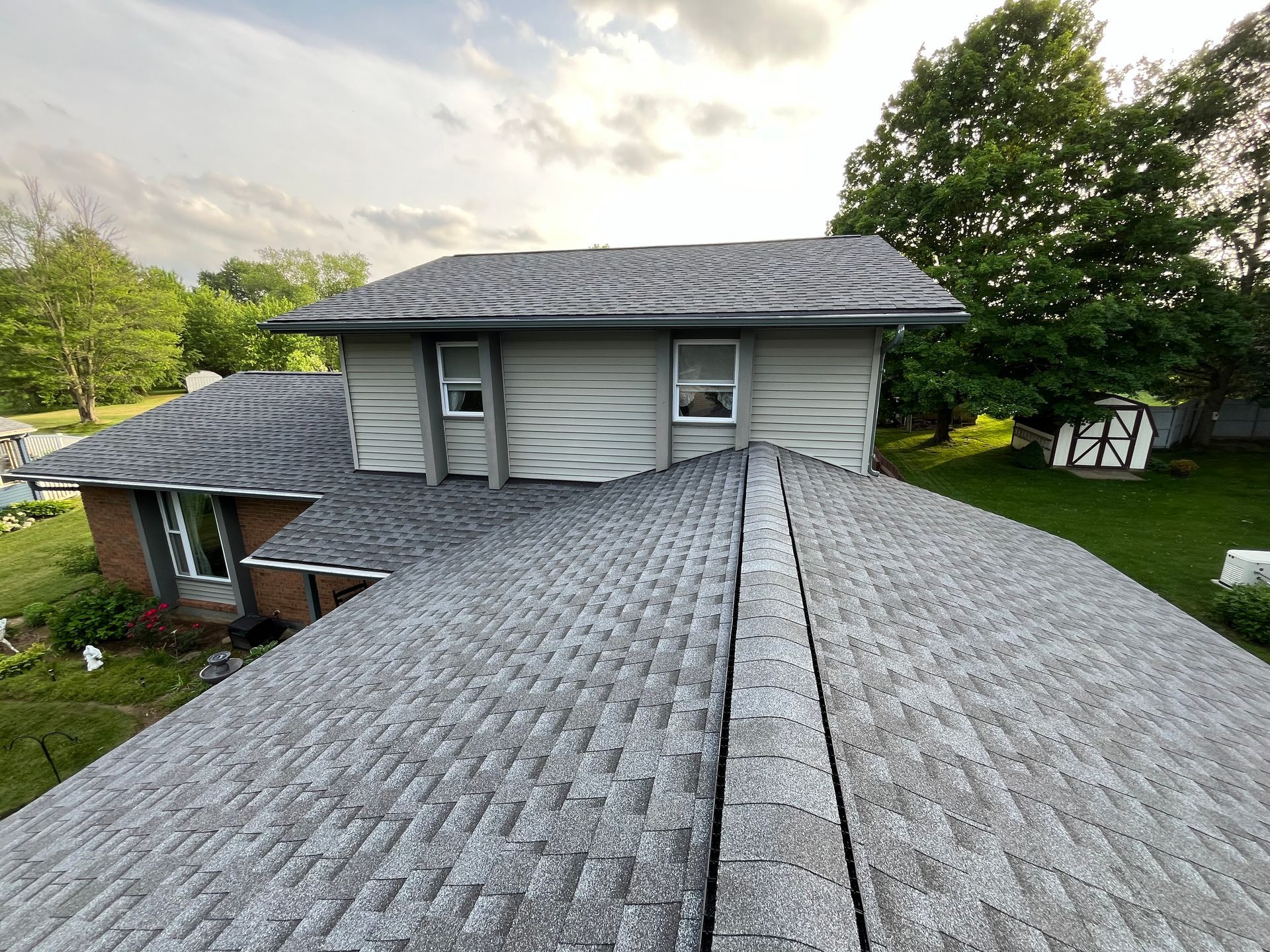Roofed house with gray shingles and a tree-lined yard under a cloudy sky
