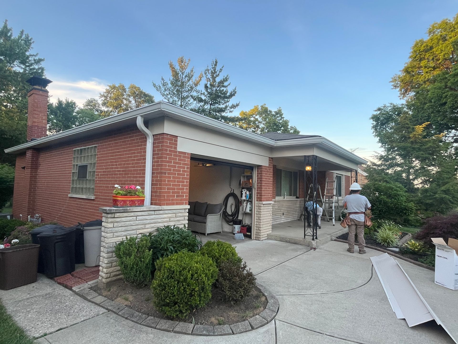 Brick house with open garage, people standing in driveway, and moving boxes outside at dusk