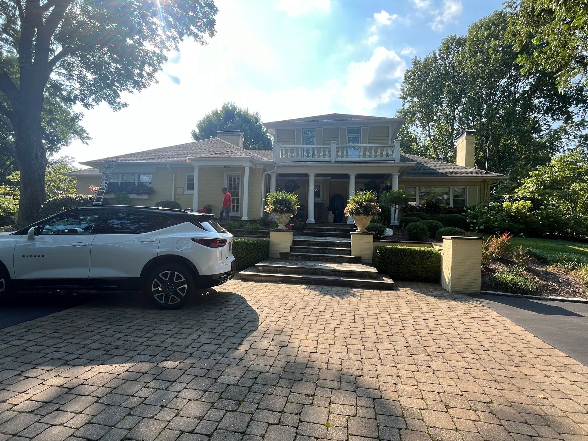 White SUV parked in front of a large brick house with a porch and stone driveway in bright sunlight