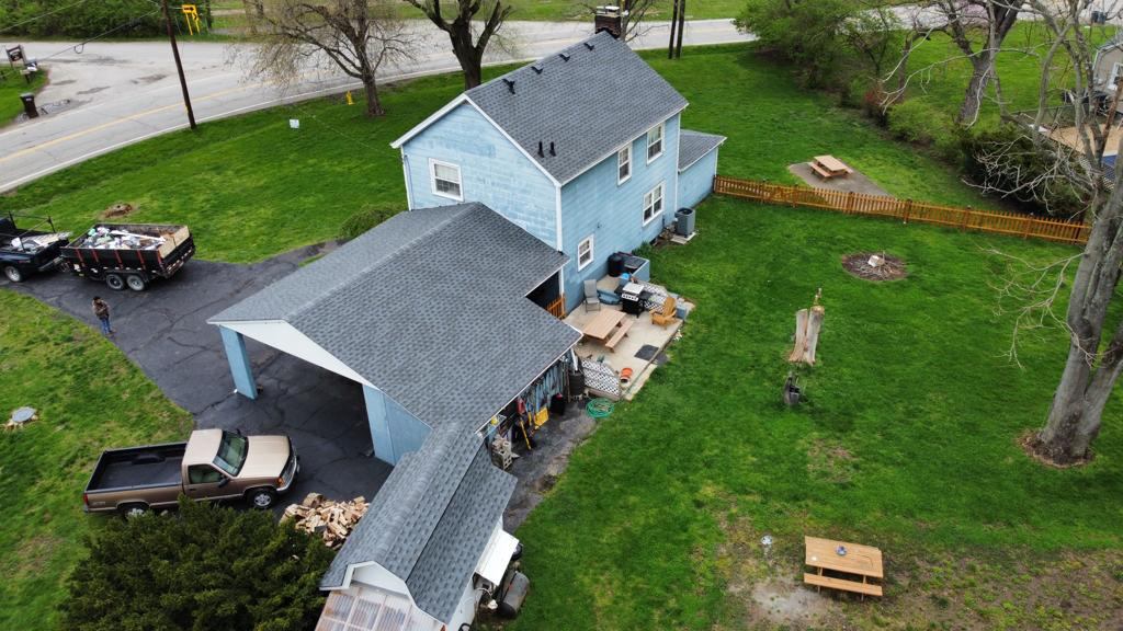Aerial view of a light blue house with a carport, patio furniture, parked cars, and yard work materials.