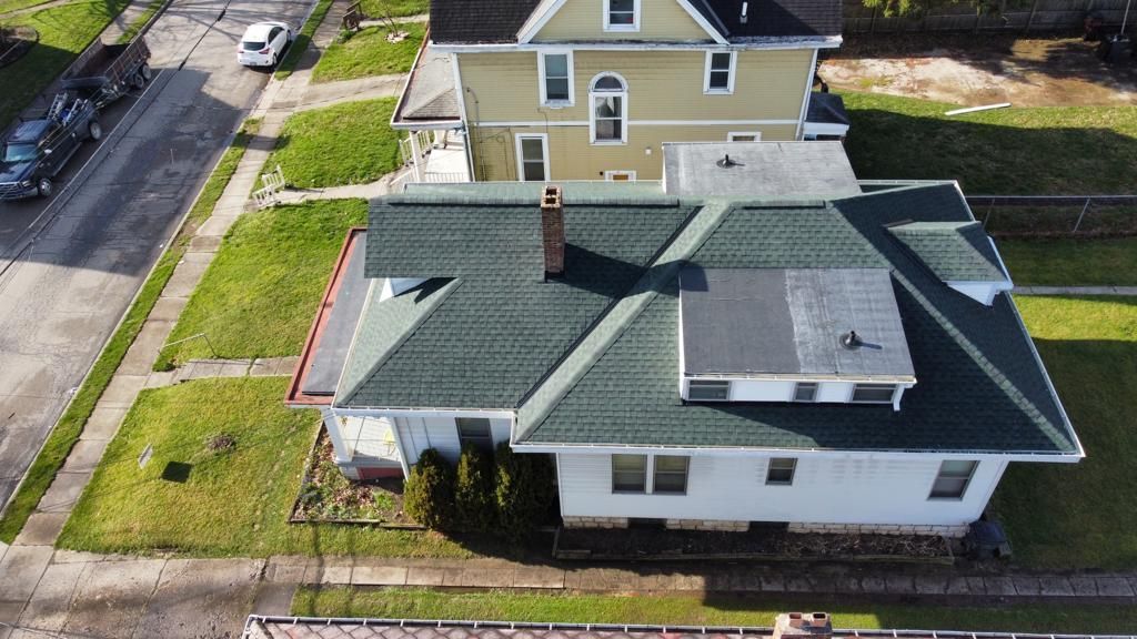 Aerial view of a white house with a green roof and a small rooftop deck beside a street and neighboring homes.