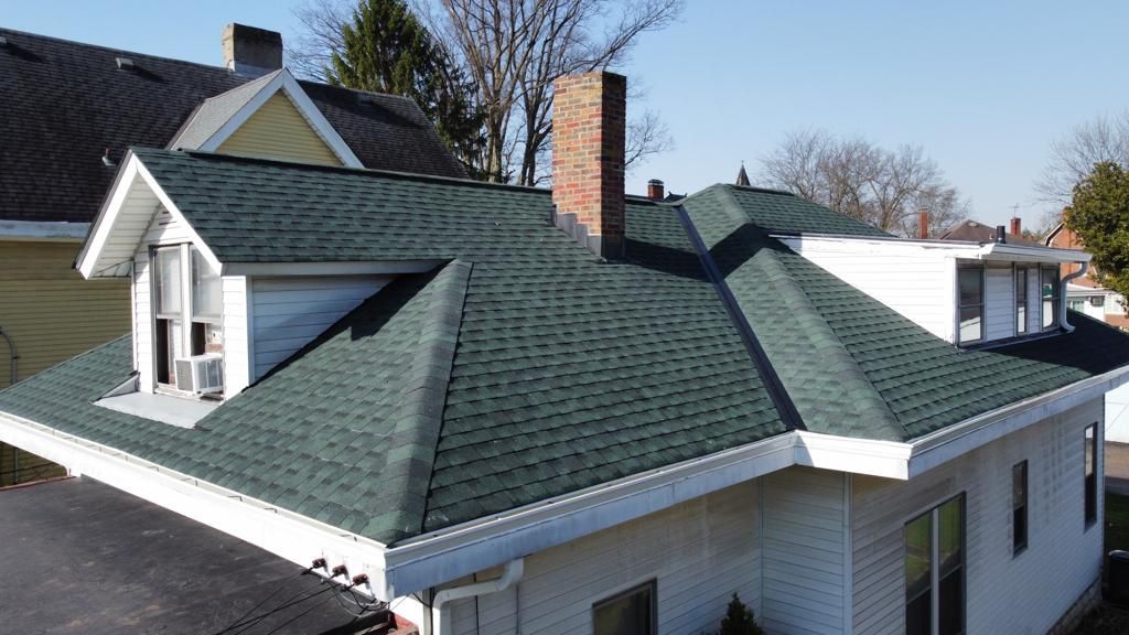 Green shingled roof with brick chimney on a white house, seen from above under a clear blue sky