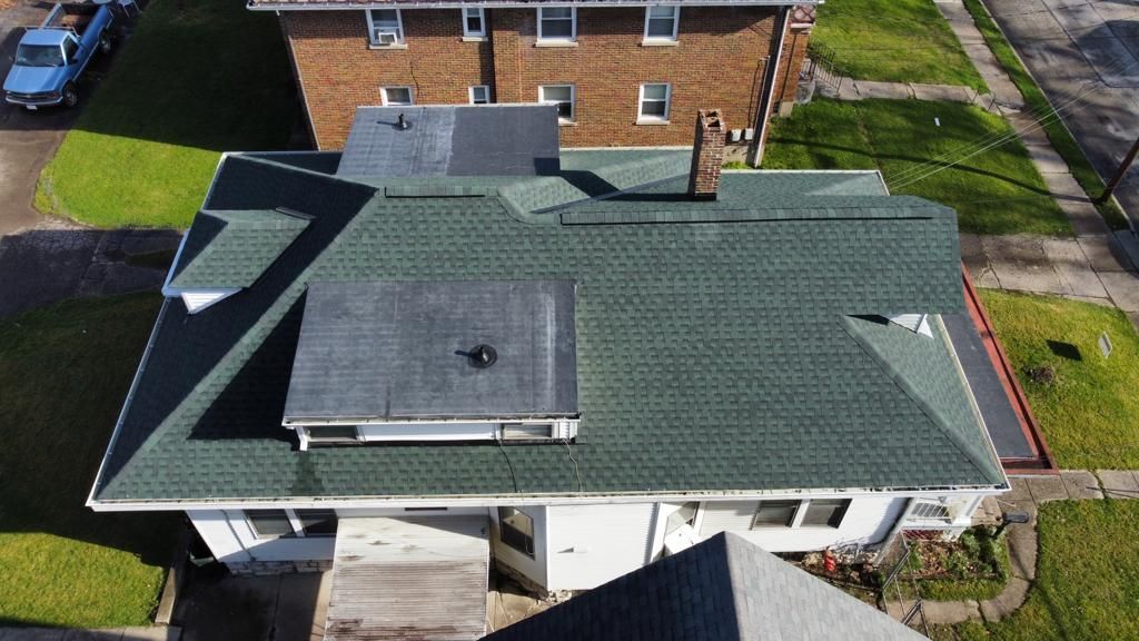 Aerial view of a gray-roofed house with solar panels and a driveway beside a brick building.