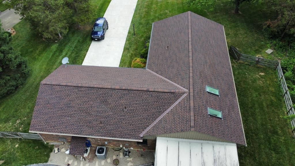 Aerial view of a brown-roofed house with a driveway, parked car, and surrounding green yard.