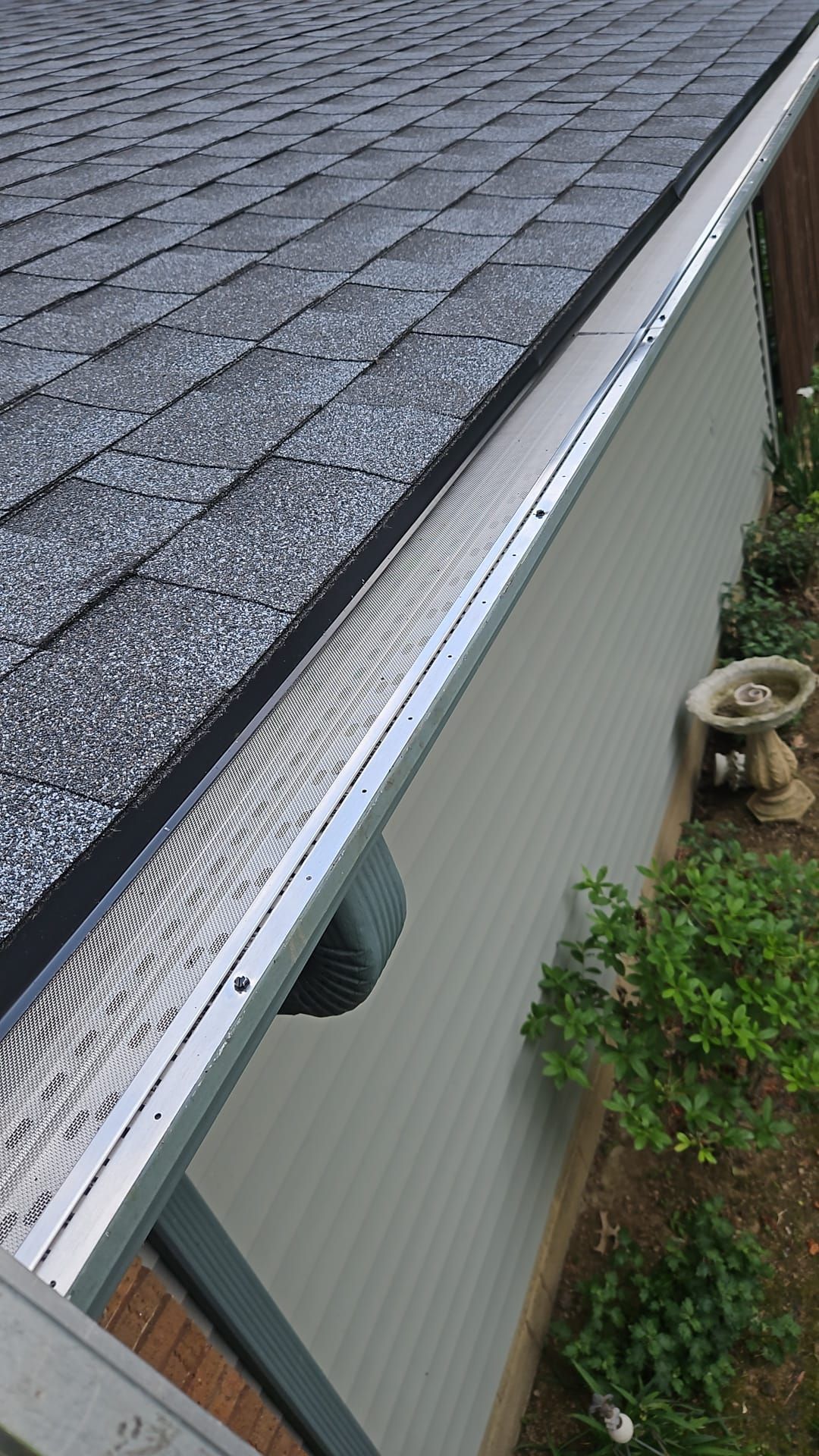 Roof gutter along gray shingle roof beside beige vinyl siding and a small garden below