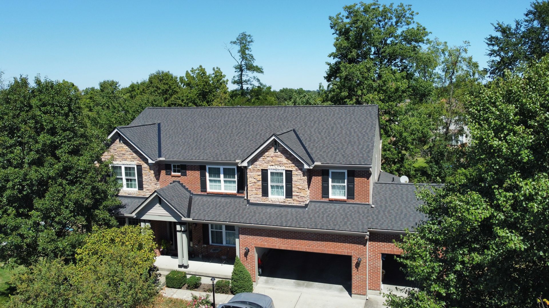 Two-story brick house with gray roof and attached garage, surrounded by dense green trees