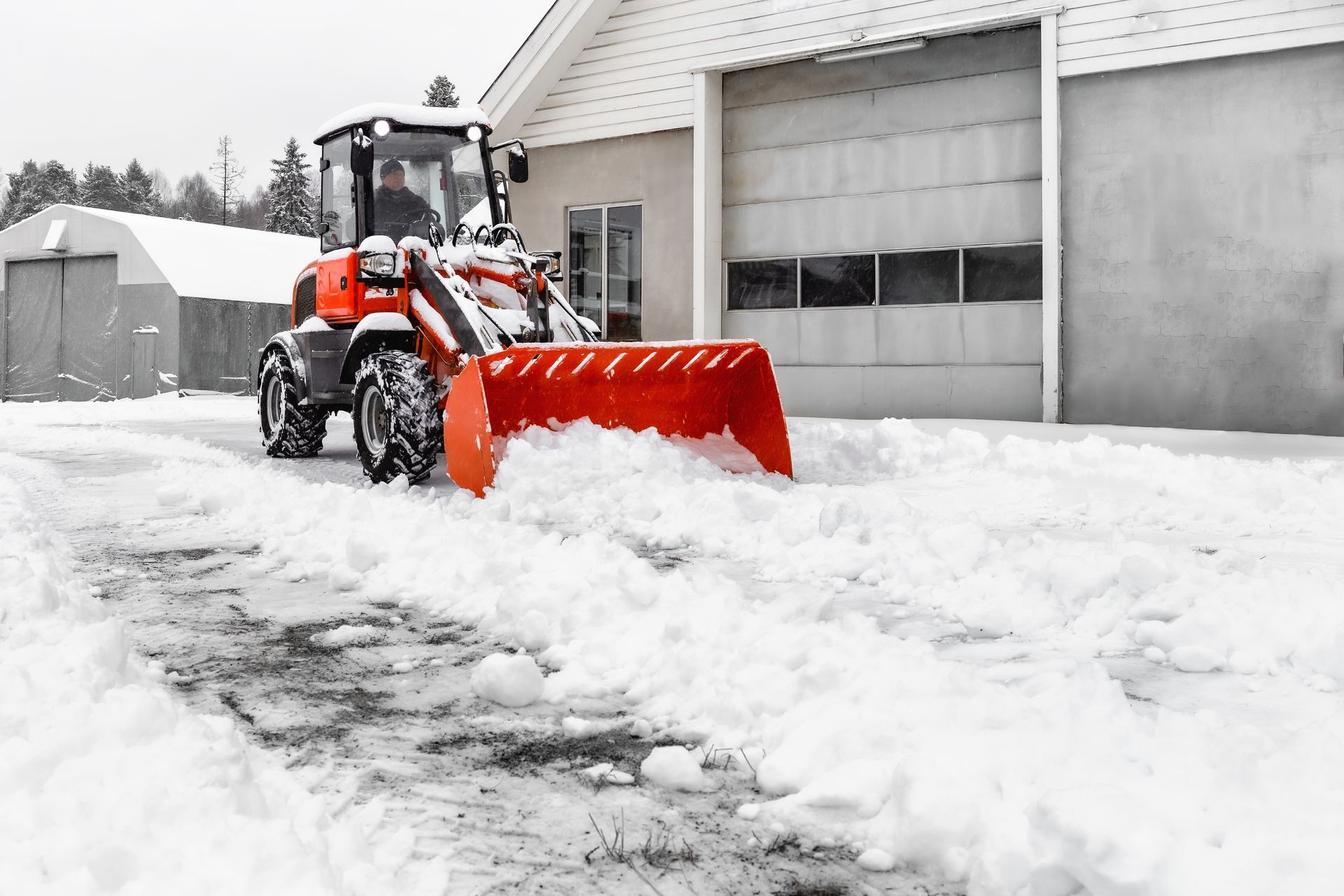 Orange snowplow clearing snow from a driveway in front of a building on a winter day.