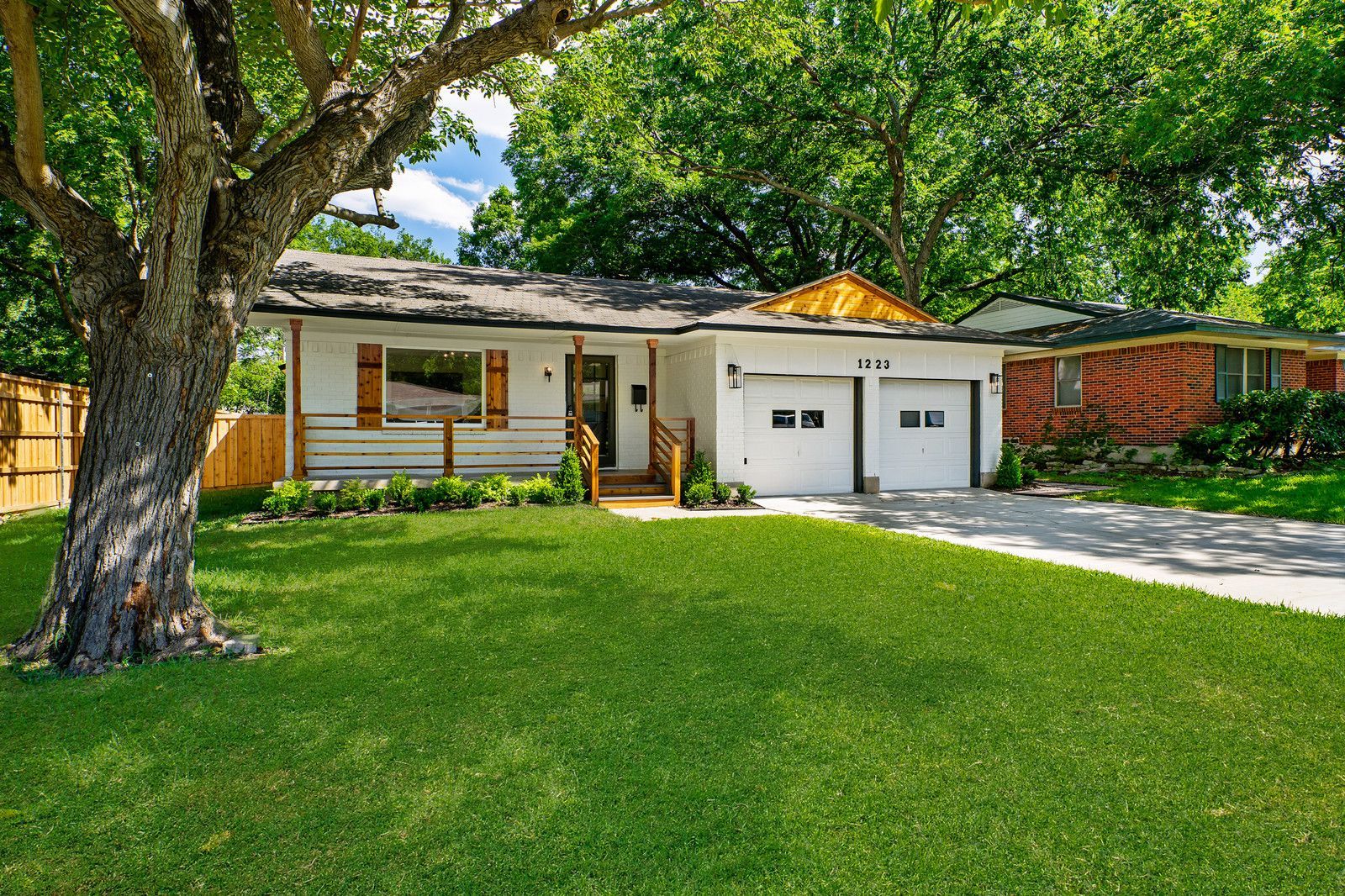 A white, single-story house with a wooden porch and attached two-car garage sits on a green lawn shaded by a large tree.