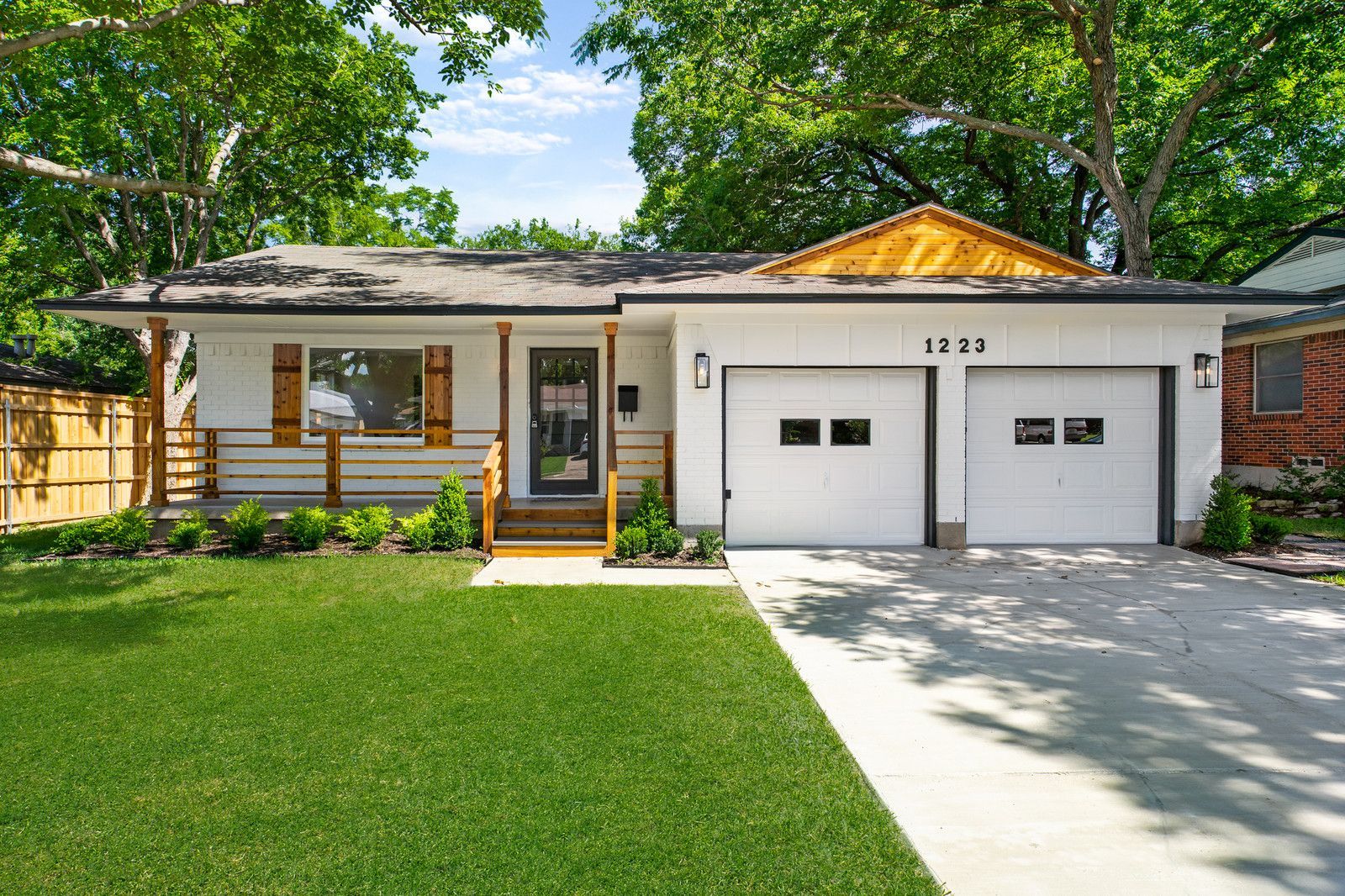 A single-story white house with a two-car garage, wooden porch accents, and a green lawn under large shade trees.
