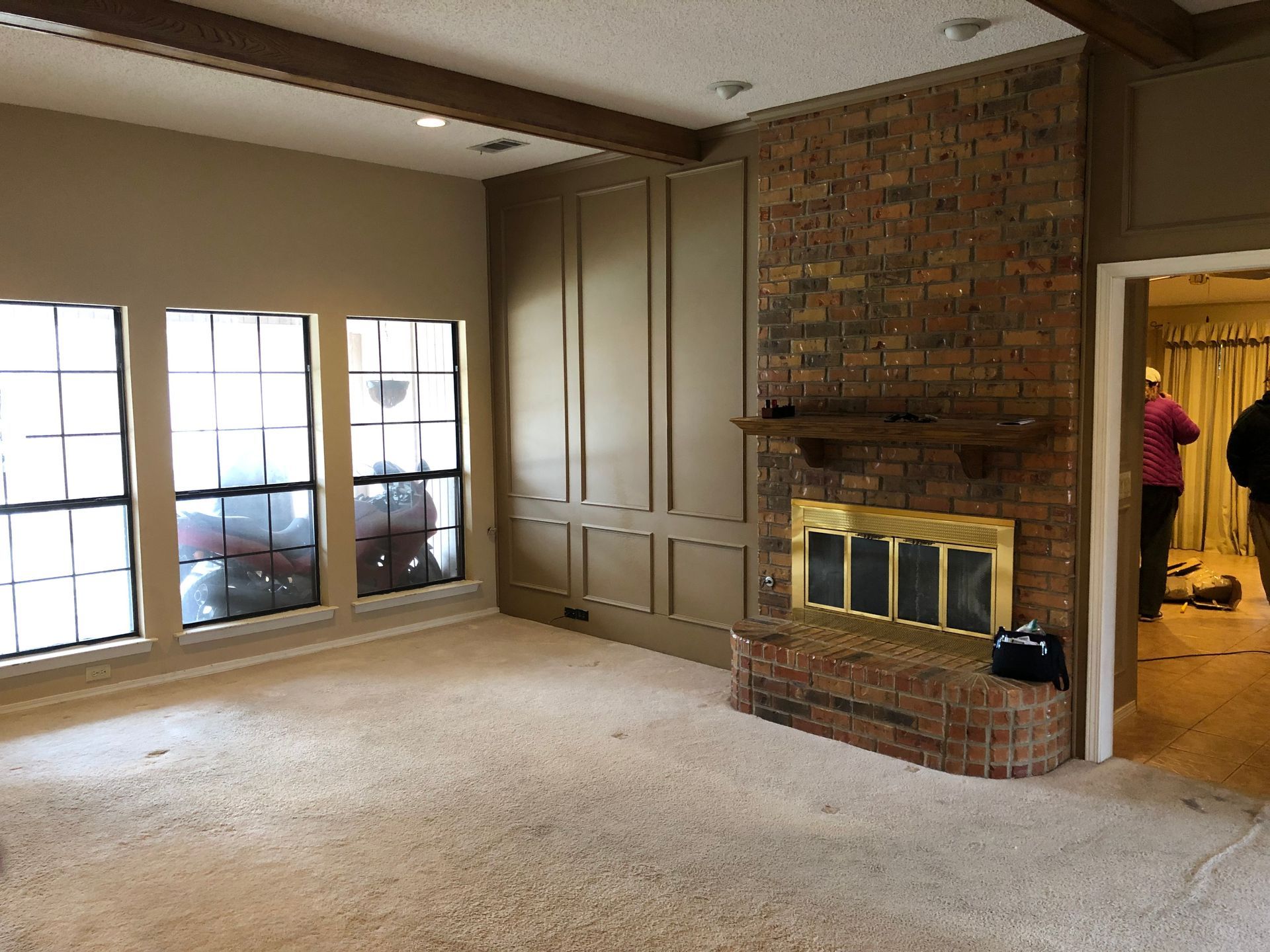 A room with windows, tan paneled walls, and a brick fireplace featuring a brass screen, seen before a doorway.