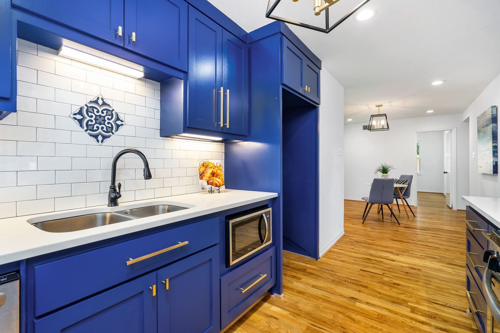 A modern kitchen with vibrant blue cabinets, a white tile backsplash, a sink, and a microwave, opening to a dining area.