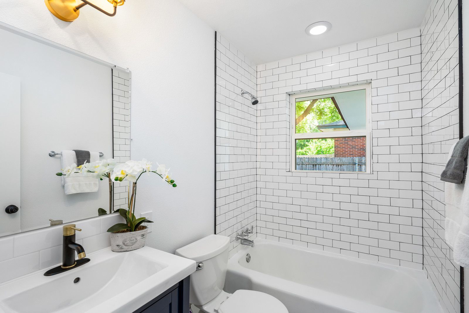 A bathroom featuring a white vanity, a tiled shower-tub area with a window, and black accents against white walls.