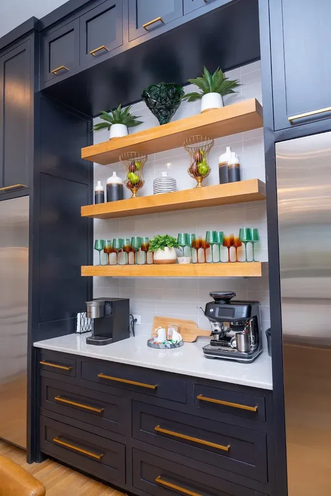 A dark blue kitchen cabinet unit with wooden open shelves displaying glassware, plants, and coffee machines on a counter.