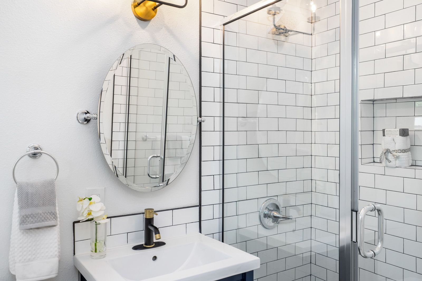 A bright bathroom with a white sink, oval mirror, wall-mounted towel ring, and a walk-in shower with white subway tiles.