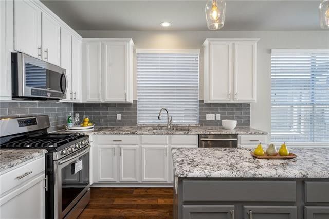 A modern kitchen featuring white cabinets, grey tiled backsplash, stone countertops, stainless steel appliances, and island.