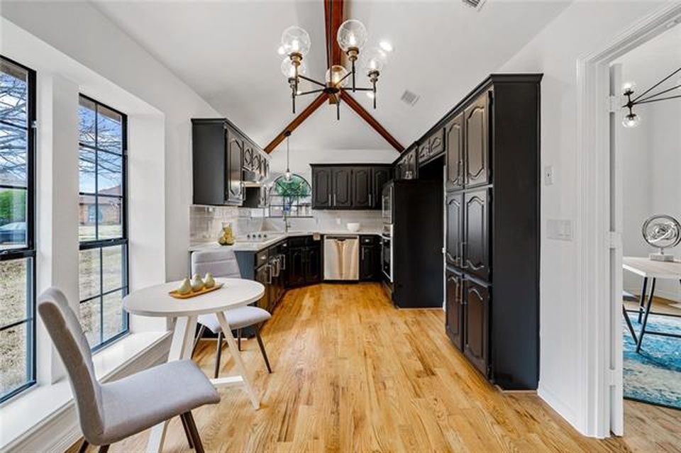 A small, bright kitchen with dark cabinets, a vaulted wood-beamed ceiling, and a light wood-topped dining table by a window.