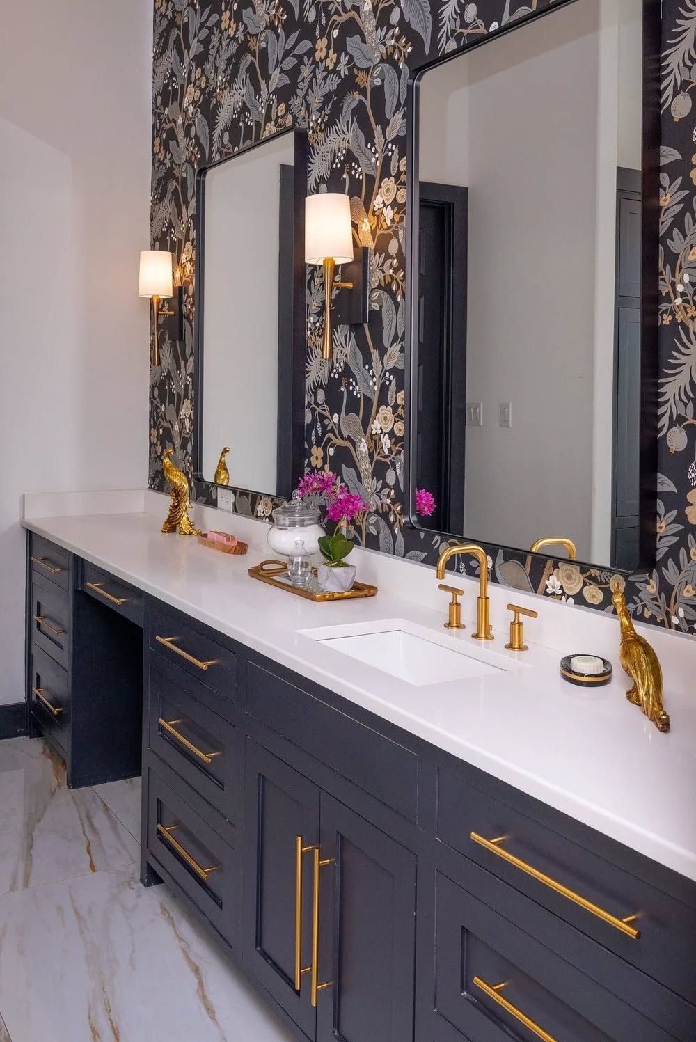 A double vanity with navy cabinets, white countertops, gold fixtures, two framed mirrors, and black floral wallpaper.