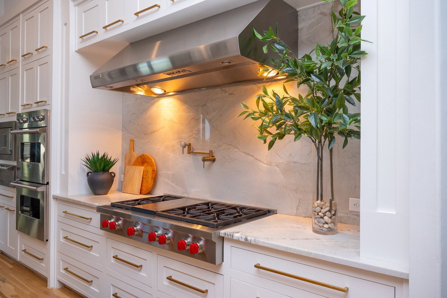 A high-end kitchen with white cabinets, gold hardware, a gas range, a stainless steel hood, and marble backsplash.
